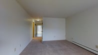 This three-floor property offers a clean, functional layout with a straightforward design. The living area visible in the image is finished with neutral gray carpeting and crisp white walls, creating a bright and versatile space ready for personalization. A baseboard heating unit runs along the exterior wall, providing efficient climate control throughout the cooler months.

A central hallway extends from the main living area, connecting to additional rooms and offering a glimpse of natural light filtering in through a glass-paneled door at the far end. The hallway is lined with warm-toned wood door frames, adding a subtle contrast to the otherwise neutral color palette. Overhead, flush-mounted ceiling lighting illuminates the corridor, ensuring visibility throughout.

The layout appears well-organized, with doorways branching off the hallway to provide access to separate rooms. The consistent carpeting flows through the living space and into the connecting areas, unifying the floor plan. The overall ambiance is clean and understated, offering a neutral canvas well-suited for a variety of interior styles. With three floors of living space, the property provides ample room across multiple levels.