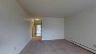 This three-floor property offers a clean, functional layout with a straightforward design. The living area visible in the image is finished with neutral gray carpeting and crisp white walls, creating a bright and versatile space ready for personalization. A baseboard heating unit runs along the exterior wall, providing efficient climate control throughout the cooler months.

A central hallway extends from the main living area, connecting to additional rooms and offering a glimpse of natural light filtering in through a glass-paneled door at the far end. The hallway is lined with warm-toned wood door frames, adding a subtle contrast to the otherwise neutral color palette. Overhead, flush-mounted ceiling lighting illuminates the corridor, ensuring visibility throughout.

The layout appears well-organized, with doorways branching off the hallway to provide access to separate rooms. The consistent carpeting flows through the living space and into the connecting areas, unifying the floor plan. The overall ambiance is clean and understated, offering a neutral canvas well-suited for a variety of interior styles. With three floors of living space, the property provides ample room across multiple levels.