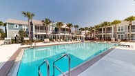 This resort-style outdoor pool area is a standout amenity designed for relaxation and recreation. The centerpiece is a generously sized swimming pool with brilliantly clear, blue-green water, accessible via stainless steel entry rails and complemented by a shallow wading ledge. Adjacent to the main pool is a dedicated spa, providing a dedicated space for relaxation. The expansive pool deck is finished in a smooth, light-colored surface and is lined with multiple sun loungers and chairs, offering ample space for sunbathing under open skies. Tall, swaying palm trees are interspersed throughout the deck, lending a tropical ambiance to the setting. A pergola-style shade structure provides a sheltered retreat from the sun. On one side of the deck, a well-appointed outdoor kitchen and bar area features built-in grills and counter seating, complemented by patio dining furniture and a market umbrella — ideal for casual outdoor gatherings. A decorative green wall panel adds a lush, landscaped accent to the space. The entire area is enclosed by an iron fence, with the surrounding residential buildings featuring balconies that overlook the pool, creating a cohesive and vibrant community atmosphere.