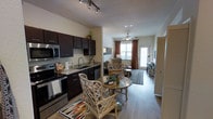 This single-floor unit showcases an open-concept layout that seamlessly connects the kitchen, dining, and living areas, creating a cohesive and functional living environment. The kitchen is outfitted with dark espresso cabinetry, granite countertops, and stainless steel appliances, including a built-in microwave, dishwasher, and range. A undermount sink with a modern faucet adds a refined touch to the workspace.

Wood-style flooring flows throughout the main living areas, lending warmth and visual continuity to the space. The dining area accommodates a round table with wicker-style chairs, positioned conveniently between the kitchen and living room. The living area is anchored by a ceiling fan and benefits from natural light streaming through large windows dressed with warm-toned drape panels.

A sliding glass door at the rear of the living room provides access to an outdoor area, enhancing the sense of openness. Track lighting in the kitchen and a ceiling fan in the living space ensure well-distributed illumination throughout. A louvered closet door near the entry adds practical storage without disrupting the overall aesthetic of the unit.
