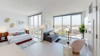 This contemporary unit is designed with a clean, modern aesthetic that emphasizes openness, natural light, and elevated city living. The open-concept living area is anchored by dramatic floor-to-ceiling windows that span two walls, offering sweeping panoramic views of the surrounding cityscape and flooding the space with natural light. Light-toned hardwood-style flooring runs throughout, enhancing the airy and cohesive feel of the interior.

The kitchen is thoughtfully designed with two-tone cabinetry — high-gloss white upper cabinets paired with warm wood-grain lower cabinets — complemented by a crisp white countertop and a full suite of stainless steel appliances including a gas range, microwave, dishwasher, and refrigerator. A central island with built-in open shelving provides additional prep space and storage, while globe pendant lights add a stylish finishing touch.

The living and sleeping areas flow seamlessly within the open layout, with the corner window configuration making the space feel expansive. A cozy dining nook positioned directly at the floor-to-ceiling windows takes full advantage of the elevated city views.

The bathroom is appointed with marble-look wall and floor tiles, a sleek floating vanity with a white countertop, a large mirror, built-in open shelving, and a soaking tub with a tiled surround — delivering a clean, spa-like atmosphere throughout.