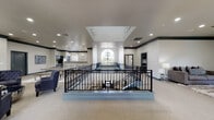 This well-appointed community facility is designed to impress, combining elegant architectural details with versatile, functional spaces across a single expansive floor. At the heart of the layout is a dramatic open mezzanine area encircled by ornate wrought iron railings with decorative scroll detailing. A soaring arched window floods this central space with natural light, complemented by a grand multi-tiered chandelier that serves as a striking focal point. A raised skylight above adds further brightness to the interior.

The full kitchen is outfitted with stainless steel appliances, gray cabinetry, white countertops, and a double-basin sink set into a generous island. Elegant pendant lighting illuminates the workspace. An adjacent bar area features a raised counter with modern acrylic barstools, additional cabinetry, and matching pendant fixtures, creating an ideal setting for social gatherings.

A dedicated billiards room offers carpeted flooring, bold dark crown molding, multiple windows with garden views, and a contemporary sputnik-style chandelier overhead. The lounge area provides plush seating, recessed lighting, and tasteful artwork. A fitness area is also visible, rounding out this multifaceted space.