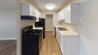 This 1-bedroom, 1-bathroom unit is laid out across a single floor, offering a functional and comfortable living environment. The living area is spacious and bright, featuring wood-style laminate flooring and a large window with vertical blinds that allows natural light to fill the room. A ceiling fan adds both style and practicality to the space. Textured ceilings and neutral-toned walls are consistent throughout, creating a clean, cohesive aesthetic.

The galley-style kitchen is thoughtfully updated with crisp white shaker cabinetry, light countertops, and wood-style flooring. Black appliances — including a gas range, refrigerator, and dishwasher — provide a striking contrast against the bright cabinetry. A stainless steel sink and ample counter space complete this efficient kitchen layout.

The bedroom is generously sized with plush carpeting, neutral walls, and a large window with vertical blinds, offering a quiet and comfortable retreat. Baseboard heating ensures warmth throughout the cooler months.

The bathroom features wood-style flooring, a white vanity, and a full tub-shower combination with white tile surround and brushed nickel fixtures, conveying a clean and updated finish throughout. This unit combines practical updates with a welcoming, move-in ready atmosphere.