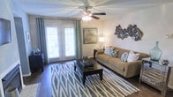 This single-story space centers around a well-appointed living room designed for both comfort and style. The room features rich hardwood flooring that anchors the space and complements the neutral, warm wall tones throughout. A ceiling fan with an integrated light fixture is positioned centrally overhead, providing both air circulation and ambient lighting.

Large French doors framed by elegant teal drape panels serve as a focal point along one wall, allowing generous natural light to fill the room while offering access to the outdoors. Horizontal blinds provide privacy and light control when needed.

The living area is furnished with a classic tan sofa accented by patterned throw pillows in coordinating blue and white tones. A dark-stained coffee table sits atop a geometric area rug in grey and white, adding visual texture to the floor. A side console table holds a table lamp, providing warm task lighting, while a framed artwork piece and a sculptural wall accent in earth tones contribute decorative interest to the walls.

Overall, the space presents a cohesive, contemporary aesthetic with a balanced mix of neutral tones, natural materials, and thoughtful decorative details.