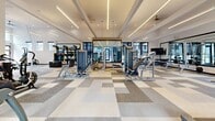 Located in Northglenn, Colorado, this fitness center offers a generously sized, single-floor exercise space designed with both functionality and aesthetics in mind. The layout is open and well-organized, providing ample room to move between equipment zones without feeling crowded.

The architecture features soaring ceilings with a coffered design accented by linear LED lighting and recessed fixtures, creating a bright and energizing atmosphere. Large windows line the upper walls, flooding the interior with natural light, while a full-panel glass garage door opens the space to outdoor views of the surrounding community, enhancing ventilation and connectivity with the exterior.

The flooring consists of a stylish checkerboard-patterned carpet tile in neutral tones of gray and white, adding visual interest while providing comfort underfoot. Ceiling fans complement the modern HVAC system to maintain a comfortable workout environment.

Equipment is thoughtfully arranged throughout the space and includes treadmills, stationary bikes, air bikes, rowing machines, ellipticals, cable machines, strength training stations, free weights, and a functional training rig with accessories. A dedicated cardio cycling area and mirrored walls further enhance the training experience. Wall-mounted TVs are positioned for visibility across multiple workout zones, rounding out this well-appointed fitness facility.