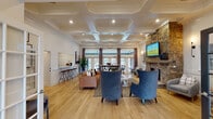 Located at 1904 Parable Way in Cary, NC, this single-story community amenity space combines refined design with practical functionality. The main lounge area serves as the centerpiece of the facility, showcasing a striking coffered ceiling with recessed lighting that adds architectural depth and a sense of grandeur. Light hardwood flooring flows throughout, complementing the bright, airy atmosphere created by large French doors and floor-to-ceiling windows that invite abundant natural light. A full-height stacked stone fireplace anchors one wall, topped with a mounted flat-screen television, while comfortable seating arrangements—including plush velvet accent chairs and a leather sofa—provide an inviting gathering space. A high-top communal table positioned near the windows offers an additional seating option. The entry corridor features curated wall art, wainscoting, and elegant pendant lighting, creating a welcoming arrival experience. A galley-style kitchenette is outfitted with dark cabinetry, white countertops, open shelving, and full appliances. The fitness center occupies a dedicated wing with vaulted, dark-painted ceilings, exposed wood beams, and ample natural light. It includes cardio equipment, strength training stations, floor-to-ceiling mirrors, and a separate stretching and functional training area with durable flooring.