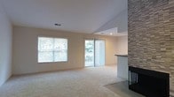 This single-floor home offers a welcoming and well-lit living space defined by clean lines and thoughtful design. The main living area features vaulted ceilings that enhance the sense of openness, while plush wall-to-wall carpeting adds warmth and comfort underfoot. Natural light floods the room through a large window fitted with horizontal blinds and a sliding glass door dressed with vertical blinds, the latter providing direct access to an outdoor deck with views of surrounding greenery. A standout architectural feature is the floor-to-ceiling stone-clad fireplace accent wall positioned to one side of the room, adding texture and a striking visual focal point. The neutral, light-toned walls complement the natural palette of the space, creating a calm and airy ambiance throughout. A recessed light in the adjoining area hints at a connected kitchen or dining space, suggesting a practical open-concept flow between living zones. The overall design balances simplicity with character, making this a versatile and inviting space suitable for a variety of living arrangements.