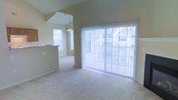 This two-floor property showcases an open and airy interior highlighted by soaring vaulted ceilings that amplify the sense of space throughout the main living area. Neutral-toned carpet flows across the floor, contributing to a clean and cohesive aesthetic. A large sliding glass door fitted with vertical blinds serves as a prominent architectural feature, flooding the living area with natural light while providing direct access to an outdoor space. Views of the surrounding exterior are visible through both the sliding door and nearby windows, enhancing the connection between indoors and outdoors. A partial wall or breakfast bar divides the living area from an adjacent kitchen or dining space, maintaining an open-concept feel while offering subtle spatial definition. A pendant light fixture hangs above the transition area, adding a functional yet decorative touch. Additional windows positioned at varying heights, including what appears to be a skylight or upper-level window, further brighten the interior. The overall ambiance is light, neutral, and versatile, providing a welcoming and functional environment throughout both levels of the home.