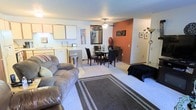 This single-floor space showcases an open-concept design that integrates the living, dining, and kitchen areas into one cohesive and functional layout. The living area is anchored by plush, neutral-toned carpeting and a large area rug with a bold border pattern, providing a comfortable and welcoming atmosphere. A generously sized sofa offers ample seating, making the space well-suited for relaxing or entertaining.

A standout feature is the warm burnt-orange accent wall that adds a distinct pop of color and visual depth to the dining area, where a dark wood dining table and matching chairs create an intimate setting. A ceiling fan with a light fixture serves as a practical focal point overhead, ensuring comfort throughout the space.

The kitchen, visible from the living area, features light wood cabinetry, white countertops, and essential appliances, maintaining a clean and efficient workspace. The open sightlines between areas enhance the sense of spaciousness throughout the unit. Decorative touches, including wall art, a large mirror, and floor lamps, contribute to a personalized and lived-in ambiance throughout the space.