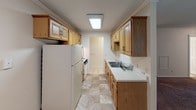 This single-floor unit offers a straightforward, functional layout across several distinct living areas. The galley-style kitchen is well-equipped with warm wood cabinetry lining both walls, white countertops, a double-basin sink, and tile flooring. A skylight-style flush fixture provides bright overhead illumination, and appliances including a refrigerator and range are included. The kitchen connects directly to the rest of the unit via an open doorway.

The living area is generously sized, featuring wall-to-wall carpet, textured ceilings, and large windows fitted with horizontal blinds that allow ample natural light. A ceiling fan with integrated lighting and a wall-mounted air conditioning unit serve the space, while baseboard heating runs along the perimeter walls. Crown molding adds a subtle finishing detail.

The bedrooms are carpeted and include natural light from windows with blinds. At least one bedroom features a wide closet with a full-width hanging rod, providing practical storage. The bathroom includes a tub-shower combination, toilet, and a vanity with a sink and mirror. Neutral, light-toned walls are consistent throughout the unit, lending a clean and cohesive feel to the overall space.