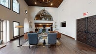 Located at 8585 La Mesa Blvd in La Mesa, San Diego, this single-floor community space showcases a striking blend of classic architectural character and modern interior design. The centerpiece of the space is a grand lounge area distinguished by dramatically soaring ceilings accented with a warm wood-paneled vaulted ceiling section and an elegant wrought-iron chandelier. Sweeping arched alcoves line the walls, adding a refined architectural detail that contributes to the space's timeless aesthetic. Rich, dark hardwood flooring flows throughout the lounge, anchoring a carefully curated seating arrangement of leather sofas and upholstered chairs arranged around a central coffee table. A mezzanine-level balcony overlooks the main lounge area, adding vertical drama and depth to the space. Large clerestory windows allow natural light to fill the upper walls, enhancing the open and airy atmosphere. Ornately carved dark wood double doors serve as a bold architectural focal point. The adjacent full kitchen is well-appointed with two-tone cabinetry in rich walnut and dark espresso finishes, white quartz countertops, stainless steel appliances including a double wall oven and refrigerator, and a central island. Decorative wall niches throughout provide tasteful display areas, adding to the space's sophisticated and welcoming ambiance.