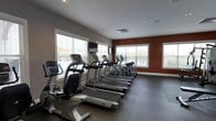 The fitness center at The Avenue is a thoughtfully designed, single-floor exercise facility built to support an active lifestyle. The space features a generous lineup of cardio equipment, including multiple treadmills and elliptical machines, as well as stationary bikes — all arranged to maximize floor space and ease of movement. The rubber flooring provides both safety and durability, making it well-suited for a high-traffic fitness environment. Natural light pours in through large, white-trimmed windows fitted with blinds, lending the space a bright and open atmosphere. The walls are painted in a neutral palette with a warm terracotta accent wall that adds a touch of character to the room. Recessed ceiling lighting supplements the natural light, ensuring the space remains well-lit at all hours. A wall-mounted television is positioned for convenient viewing during workouts. The overall layout is clean, organized, and functional, reflecting a commitment to providing residents of The Avenue with a quality fitness experience in a comfortable and well-maintained environment.