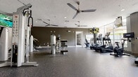 Located at 100 Myrtle St in Longwood, Florida, this fitness center offers a spacious, single-floor layout designed to support a comprehensive workout experience. The facility is outfitted with durable rubber flooring throughout, providing a safe and resilient surface suited for a wide range of exercise activities.

The cardio zone features multiple treadmills, elliptical trainers, and a stationary bike, all positioned along a wall of large floor-to-ceiling windows that flood the space with natural light and offer views of the surrounding outdoor area, including lush palm trees. Wall-mounted flat-screen televisions are positioned above the equipment for entertainment during workouts.

A full-length wall mirror runs along one side of the room, visually expanding the space and providing users with clear sightlines for form correction. The strength training area includes a multi-station cable machine, a functional trainer, and a dumbbell rack with a range of free weights, as well as stability balls for flexibility and core work. Colorful wall art adds a subtle decorative touch to this zone.

Multiple ceiling fans are mounted throughout the space, complementing the recessed lighting to maintain a comfortable environment. The overall ambiance is modern and clean, with neutral tones on the walls contributing to a focused and motivating atmosphere.