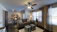 This single-floor residence offers a thoughtfully designed open-concept layout that seamlessly connects the living and dining areas into one cohesive, functional space. The living area is anchored by a plush gray sofa accented with decorative throw pillows and complemented by a pair of striped upholstered ottomans resting atop a coordinating area rug. A ceiling fan with integrated lighting fixture serves as a central overhead element, providing both comfort and illumination. Track lighting along the ceiling adds an additional layer of ambient light, enhancing the modern aesthetic of the space. Large windows dressed with sheer white curtains and grommet-top draping panels invite generous natural light while maintaining privacy. Dark hardwood-style flooring runs throughout, providing a rich contrast to the neutral-toned walls. The dining area, visible in the background, features bar-height seating and contemporary pendant-style track lighting overhead, ideal for casual meals or entertaining. A framed abstract landscape painting adorns the wall, adding a warm artistic touch. The overall ambiance is clean, modern, and inviting, with a cohesive color palette of grays, whites, and muted yellows that creates a comfortable and stylish living environment.
