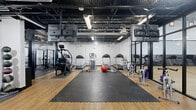 Located at 100 Hawk Ridge Dr in Iowa City, Iowa, this fitness center offers a comprehensive and well-organized workout facility across a single, expansive floor. The space is designed with an industrial aesthetic, featuring an exposed ceiling with visible ductwork, steel beams, and grid-style framing that lends an open, warehouse-like feel. Bright overhead lighting ensures the entire area is well illuminated throughout.

The facility is divided into distinct training zones. A dedicated cardio area houses multiple treadmills, elliptical trainers, a StairMaster, and a rowing machine, all arranged on interlocking rubber mat flooring with wood-look flooring alongside. Stability balls and a BOSU ball are also available in this zone.

A strength training area features multiple power racks and squat stations set on durable rubber flooring, providing ample space for barbell work. An adjoining free weights section includes a full rack of dumbbells, kettlebells, weight plates, and adjustable benches, with large floor-to-ceiling mirrors — branded with Iowa Hawkeyes signage — lining the walls to maximize the sense of space and allow for form monitoring.

The facility also includes wall-mounted televisions, a water fountain station, and clearly marked entry and exit doors. The combination of varied flooring surfaces, structured equipment zones, and industrial design creates a functional and motivating training environment.