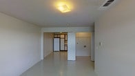 This single-floor space presents an open, minimalist layout characterized by white walls and smooth, light-toned flooring that reflects ambient light throughout. The main living area is spacious and unfurnished, offering a blank canvas well-suited to a variety of furniture arrangements. A flush ceiling light fixture with a warm-toned, geometric design serves as a subtle focal point overhead, complementing the otherwise neutral palette.

Visible from the main area, the kitchen is partially separated by a white partition wall, creating a defined yet connected zone. The kitchen features a white refrigerator, cabinetry with warm wood-toned accents, and what appears to be a countertop workspace. The cabinetry design introduces a touch of contrast against the predominantly white interior. An additional open area is accessible beyond the partition on the right side, suggesting further usable space within the unit.

The overall ambiance is clean, bright, and straightforward, with a functional design aesthetic. The light flooring and white surfaces work together to maximize the sense of openness throughout the space, making it feel airy and easy to customize.