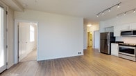 This single-floor unit offers a clean, modern design with an open-concept layout connecting the living area and kitchen. The living space features light hardwood-style flooring, crisp white walls, and track lighting overhead, creating a bright and functional atmosphere. Large windows with vertical blinds allow generous natural light to fill the room, while a wall-mounted HVAC unit provides climate control.

The kitchen is well-appointed with white cabinetry, dark countertops, and a full suite of stainless steel appliances, including a refrigerator, range, over-the-range microwave, and dishwasher. A window above the sink adds additional natural light to the workspace.

The bedroom is carpeted for comfort and features a large window with outdoor views of surrounding greenery, along with a baseboard heater. The space is quiet and well-lit, offering a peaceful retreat.

The bathroom includes white cabinetry with brushed nickel hardware, a dark countertop with an undermount sink, and hardwood-style flooring that continues from the main living areas. The layout is practical and well-organized, with a large mirror and stylish light fixtures above the vanity. Overall, the unit presents a cohesive, contemporary aesthetic throughout.