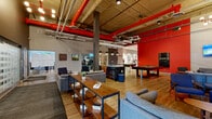 Located at 233 Kennedy St in Winnipeg, this generously sized amenity space offers a dynamic blend of industrial design and modern comfort across a single open floor. The main lounge area is defined by soaring exposed concrete ceilings with striking red-painted ductwork and piping, concrete support columns, and warm hardwood-style flooring that runs throughout. Bold red accent walls add energy and visual contrast to the space, while multiple seating arrangements featuring contemporary grey sofas, mid-century-style armchairs, and open wood-and-metal shelving units create distinct social zones within the open-plan layout. A standout floor-to-ceiling black-and-white cityscape mural serves as a dramatic focal point in one lounge area, complemented by a wall-mounted flat-screen television. A central wet bar with a dark stone countertop and pendant lighting provides a functional gathering point, while a full-sized pool table anchors the recreation zone. Original artwork and framed architectural photography are displayed throughout, adding a curated, gallery-like quality. The hallway connects to additional amenities, including package lockers and restrooms, and is finished with light wood flooring, recessed lighting, and continued red wall accents, maintaining a cohesive aesthetic throughout the entire space.