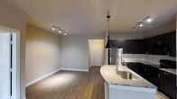 This single-floor space in Carrollton offers a thoughtfully designed open-concept layout that seamlessly connects the kitchen and living areas. The kitchen is a standout feature, showcasing dark espresso cabinetry, granite countertops with a curved breakfast bar, a stainless steel sink, and stainless steel appliances including a refrigerator. A sleek black pendant light hangs above the kitchen island, adding a modern touch, while track lighting illuminates both the kitchen and living areas with precision and style. The living area is open and inviting, with warm hardwood-style flooring that runs throughout the space, creating a cohesive and welcoming atmosphere. Neutral gray walls provide a versatile backdrop suited to a variety of décor styles. A white paneled door leads to a private entryway, contributing to the unit's clean, contemporary aesthetic. The combination of modern lighting, quality finishes, and an open floor plan makes this Carrollton space an excellent blend of functionality and style.