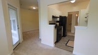 This single-floor space is designed with an open-concept layout that connects the kitchen and living area, creating a cohesive and functional flow throughout. The kitchen is equipped with black appliances, including a gas range and a full-sized refrigerator, complemented by white cabinetry that provides ample storage. A partial dividing wall with a breakfast bar-style peninsula separates the kitchen from the adjoining living space, adding definition without sacrificing openness. The kitchen features wood-look flooring, which transitions smoothly into the plush carpeting of the living area. Flush-mount ceiling lighting illuminates both spaces evenly. Crown molding details along the upper walls add a subtle architectural refinement to the interior. A hallway visible beyond the kitchen leads to additional rooms, suggesting a practical and well-organized floor plan. The neutral color palette throughout — including warm beige walls and light-toned carpet — creates a clean, welcoming ambiance. The overall design balances functionality with a comfortable residential aesthetic, making efficient use of the available space on a single level.
