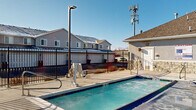 This single-level outdoor amenity complex serves as a central gathering hub for its residential community. At its heart is a generously sized swimming pool with tiled blue edges and stainless steel entry rails, complemented by a separate hot tub nearby. The pool deck is finished in smooth concrete, providing ample space for lounging and socializing, and is equipped with outdoor seating including patio tables with umbrellas.

The area is enclosed by a sturdy wrought iron fence fitted with a safety ring buoy, ensuring a secure environment. Dedicated lighting poles illuminate the space for evening use, while an ADA-accessible ramp provides inclusive entry to the pool facilities.

Adjacent to the pool sits a well-maintained clubhouse building featuring stone accent detailing, a pitched shingled roof, and large windows that overlook the deck area. Beyond the pool enclosure, a grassy recreation area with a children's playground is visible, offering additional outdoor enjoyment.

The surrounding landscape opens up to sweeping views of snow-capped mountain ranges under wide, clear skies, creating a striking natural backdrop. The overall atmosphere of this amenity space is open, functional, and community-oriented, designed to serve residents across all seasons.