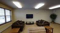 This single-floor space is designed as a comfortable communal lounge area, offering a relaxed and functional environment for gathering and leisure. The room features a neutral color palette with warm beige walls and light carpeted flooring that contribute to an inviting, understated aesthetic. A textured ceiling hosts two large rectangular flush-mounted panel lights, providing bright, even illumination throughout the space.

The focal point of the lounge is a wall-mounted flat-screen television, positioned at an ideal viewing height and complemented by a sleek glass-and-metal media stand beneath it. Seating is generous and well-arranged, with two dark brown upholstered sofas flanking the television and accented with patterned throw pillows. A lighter-toned sofa positioned in the foreground further enhances the seating capacity, making the room well-suited for group use. Wooden side tables are placed conveniently within reach of the seating areas.

The overall layout is open and thoughtfully organized to encourage conversation and shared viewing experiences. The space strikes a balance between practicality and comfort, making it a versatile lounge well-suited for relaxation and social interaction.