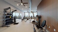 This modern fitness center is designed to accommodate a wide range of workout styles and fitness goals across a thoughtfully organized, single-floor layout. The main workout area features soaring ceilings with exposed industrial-style ductwork and ceiling fans that lend an open, energetic atmosphere. Large windows along the perimeter flood the space with natural light, while recessed lighting ensures consistent illumination throughout. The main floor is outfitted with an extensive lineup of Hoist strength machines, a dedicated free weight section with a full dumbbell rack, a cable system, adjustable benches, and plyo boxes — providing everything needed for a complete strength training routine. A row of True treadmills lines the windowed wall, offering a bright cardio experience. A separate cardio nook houses ellipticals and upright bikes, complete with a wall-mounted flat-screen TV. The dedicated cycling studio features a hardwood-style floor, spin bikes, full-wall mirrors, framed artwork, and its own mounted TV, creating an immersive environment for cycling sessions. An adjacent glass-door room appears to serve as a fitness consultation or lounge area with seating and dual monitors. Neutral gray tones, warm accent walls with geometric paneling, and curated artwork tie the entire facility together with a polished, professional aesthetic.