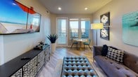 The Catalina is a thoughtfully designed single-story residence offering one bedroom and one bathroom across a well-appointed, open-concept floor plan. Light-toned luxury vinyl plank flooring flows throughout the main living areas, creating a cohesive and contemporary aesthetic.

The living area serves as the centerpiece of the home, featuring recessed lighting, a wall-mounted television, and direct access to a private balcony through glass-paneled doors that invite natural light and outdoor views into the space. A compact dining area adjacent to the living room accommodates intimate seating for two.

The kitchen is well-equipped with crisp white shaker-style cabinetry extending to the upper walls for ample storage, light countertops, and a full suite of stainless steel appliances including a gas range, over-the-range microwave, and refrigerator. The layout maximizes functionality within the space.

The bedroom offers a calm, neutral palette with plush carpet flooring, a tray ceiling accented by a ceiling fan, and a window with access to the balcony, allowing for natural light throughout the day.

The bathroom features a white vanity with a single sink, a large mirror, and tile flooring, maintaining the clean, modern design language found throughout the residence.