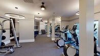 This fitness center is a thoughtfully designed, single-floor facility offering a comprehensive workout environment for users of all fitness levels. The space is divided into distinct functional zones, each outfitted with professional-grade equipment. The cardio area features multiple elliptical trainers and stationary bikes, as well as treadmills positioned near windows that allow natural light to filter in. A dedicated strength training section includes cable machines and weight benches, while a free weights zone provides a full dumbbell rack and kettlebells neatly organized on a dedicated bench with built-in shelving and navy blue cabinetry below. A stretching and functional training area is defined by large, dark-framed floor-to-ceiling mirrors that line the walls, enhancing the sense of openness and allowing users to monitor their form. Structural columns are integrated cleanly into the layout, complementing the neutral palette of warm white walls, grey carpeting, and dark accent zones. Overhead, distinctive circular ring pendant lights provide even, modern illumination throughout. A water fountain station is conveniently located within the free weights area. The overall ambiance is polished and motivating, combining a clean aesthetic with practical, well-organized equipment placement.