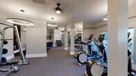 This fitness center is a thoughtfully designed, single-floor facility offering a comprehensive workout environment for users of all fitness levels. The space is divided into distinct functional zones, each outfitted with professional-grade equipment. The cardio area features multiple elliptical trainers and stationary bikes, as well as treadmills positioned near windows that allow natural light to filter in. A dedicated strength training section includes cable machines and weight benches, while a free weights zone provides a full dumbbell rack and kettlebells neatly organized on a dedicated bench with built-in shelving and navy blue cabinetry below. A stretching and functional training area is defined by large, dark-framed floor-to-ceiling mirrors that line the walls, enhancing the sense of openness and allowing users to monitor their form. Structural columns are integrated cleanly into the layout, complementing the neutral palette of warm white walls, grey carpeting, and dark accent zones. Overhead, distinctive circular ring pendant lights provide even, modern illumination throughout. A water fountain station is conveniently located within the free weights area. The overall ambiance is polished and motivating, combining a clean aesthetic with practical, well-organized equipment placement.