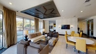 This clubroom in Houston, TX offers a thoughtfully designed, single-level communal space that balances comfort and functionality across several distinct zones. The lounge area immediately draws attention with its striking geometric coffered ceiling — a dark-toned, angular design element that anchors the room and adds architectural depth. Large floor-to-ceiling windows line the perimeter, flooding the space with natural light and providing open views of the surrounding greenery and streetscape. Light tile flooring complements the neutral palette, while plush upholstered sofas, armchairs, tufted ottomans, and glass-topped coffee tables create an inviting atmosphere for casual gatherings.

Adjacent to the lounge is a dining area featuring a round white table surrounded by mustard-yellow upholstered chairs, with a wall-mounted flat-screen television and sleek sconce lighting overhead. Dark hardwood-style flooring transitions the space into a fully equipped kitchen, showcasing rich espresso cabinetry, a mosaic tile backsplash, stainless steel appliances including a side-by-side refrigerator and dishwasher, and a generous white waterfall-edge island with leather bar seating. Pendant lighting adds warmth above the island. A separate workstation area with computers provides a functional space for productivity, completing this well-appointed communal retreat.