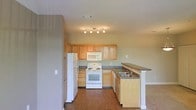 This single-floor residence offers a well-organized and functional layout designed for comfortable everyday living. The kitchen serves as a central highlight, featuring light oak wood cabinetry that provides generous storage space across both upper and lower cabinets. White appliances, including a freestanding electric range with overhead microwave, a full-size refrigerator, and a dishwasher, are neatly integrated into the layout. A peninsula with a dark laminate countertop extends from the cabinetry, housing a double-basin sink and offering additional workspace as well as a natural divider between the kitchen and the adjoining living area. The flooring throughout the kitchen and connected spaces consists of warm hardwood-style planks that add a grounded, cohesive feel to the interior. Walls are finished in a soft, neutral green-gray tone that complements the cabinetry and keeps the atmosphere light and airy. A track lighting fixture mounted to the ceiling provides focused illumination across the kitchen workspace. The open sightlines between the kitchen and living area contribute to an interconnected, spacious feel, making the layout ideal for both everyday use and casual entertaining.