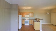 This single-floor residence offers a well-organized and functional layout designed for comfortable everyday living. The kitchen serves as a central highlight, featuring light oak wood cabinetry that provides generous storage space across both upper and lower cabinets. White appliances, including a freestanding electric range with overhead microwave, a full-size refrigerator, and a dishwasher, are neatly integrated into the layout. A peninsula with a dark laminate countertop extends from the cabinetry, housing a double-basin sink and offering additional workspace as well as a natural divider between the kitchen and the adjoining living area. The flooring throughout the kitchen and connected spaces consists of warm hardwood-style planks that add a grounded, cohesive feel to the interior. Walls are finished in a soft, neutral green-gray tone that complements the cabinetry and keeps the atmosphere light and airy. A track lighting fixture mounted to the ceiling provides focused illumination across the kitchen workspace. The open sightlines between the kitchen and living area contribute to an interconnected, spacious feel, making the layout ideal for both everyday use and casual entertaining.