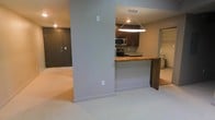 This single-floor space showcases an open-concept design that seamlessly connects the living area and kitchen. The layout is anchored by a striking live-edge wood breakfast bar, which doubles as a natural focal point and casual dining surface. A geometric pendant light hangs above, providing warm, focused illumination over the kitchen area.

The kitchen is equipped with stainless steel appliances, including a microwave and range, and features dark wood cabinetry paired with granite-style countertops. A structural column partially divides the kitchen from the main living area, adding architectural definition without closing off the space.

The living area is finished with light-toned carpeting, contributing to a soft, neutral ambiance throughout. The walls are painted in a cool gray tone, complementing the overall modern aesthetic. Recessed lighting in the living area provides additional illumination. A solid entry door with a peephole is visible at the far end, and what appears to be a utility closet is accessible nearby. The overall design balances functional living with contemporary finishes.