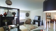 This single-floor residence offers a comfortable and thoughtfully arranged living space designed with a neutral color palette that creates a calm, welcoming ambiance. The living area serves as the heart of the home, featuring light-toned walls and carpeted flooring that adds warmth underfoot. A large window fitted with horizontal blinds allows natural light to fill the room, brightening the space and creating an open, airy feel. A decorative bookshelf unit acts as a stylish room divider, adding both function and visual interest to the layout. The living area is furnished with a rich brown leather armchair, a media console with a flat-screen television, and an ornate area rug with circular patterns that anchors the seating arrangement. Decorative touches such as a textured wall medallion, tall floor vases with branches, and carefully placed greenery add character and depth to the interior. The overall design aesthetic blends contemporary and transitional styles, resulting in a space that feels both practical and inviting. The single-floor layout ensures ease of movement and accessibility throughout, making this a well-suited space for comfortable everyday living.