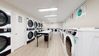 This commercial laundromat is a clean, efficiently organized single-floor facility designed to serve a high volume of customers. The space is divided into distinct functional zones, all unified by light wood-look flooring and bright white walls that contribute to a fresh, sanitary atmosphere throughout.

The main laundry area features an extensive row of Hercules-branded front-load washing machines, each clearly numbered for easy identification. Opposite the washers, stacked ADC commercial dryers line the wall, offering ample drying capacity. Instructional signage is posted above the machines to guide users through proper operation.

A central folding table runs the length of the main room, providing generous workspace for sorting and folding laundry. A utility laundry cart is also available for customer convenience. The facility incorporates a modern digital payment kiosk, allowing customers to pay with a smartphone or card, streamlining the experience.

A dedicated waiting area features a row of chairs along the wall, offering a comfortable place to relax. A wall-mounted flat-screen television is positioned above additional washers near the entrance, providing entertainment while customers wait. Natural light filters in through a window near the entry, brightening the space. Overhead fluorescent panel lighting ensures consistent, even illumination throughout the entire facility.