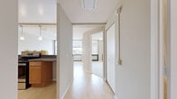 This single-floor unit offers a clean, functional layout with a neutral color palette and light hardwood flooring that runs throughout the main living areas. A central hallway connects the key spaces, providing smooth flow between rooms. The kitchen is well-appointed with warm honey-toned wood cabinetry, dark granite-style countertops, stainless steel appliances including a refrigerator and dishwasher, and pendant lighting overhead — creating a practical yet inviting cooking space. The bedroom features plush carpeting and direct access to the rest of the unit, offering a quiet retreat. The bathroom includes a wood-vanity with ample under-sink storage, a mirrored medicine cabinet, a tiled tub and shower surround, and classic white fixtures. Large windows with horizontal blinds fill the living areas with natural light and frame expansive urban views of the surrounding cityscape and skyline — a standout feature of this space. Textured ceilings are consistent throughout, and the overall design emphasizes simplicity and practicality. The open sightlines from the hallway through to the main living area enhance the sense of space and airflow, making this a comfortable and well-connected unit.