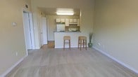 This single-floor residential space showcases an open-concept design that seamlessly connects the living area to the kitchen. The living space is finished with neutral beige carpeting, lending a warm and understated feel to the room, while the kitchen and adjoining areas transition to smooth hardwood-style flooring. Walls are painted in a soft, muted green-beige tone that creates a calm and cohesive atmosphere throughout.

The kitchen is positioned at the rear of the open layout and features white cabinetry offering ample storage, a white refrigerator, and a dark countertop that wraps around a peninsula-style breakfast bar. The bar is complemented by two wicker-style barstools, adding a casual, functional dining option. An overhead fluorescent light fixture illuminates the kitchen workspace effectively.

A doorway visible along the left wall leads to additional areas of the unit, accompanied by what appears to be a closet with a white bifold door, suggesting convenient storage access near the main living space. The overall design is clean, practical, and move-in ready, with a straightforward layout that maximizes the usable living area.