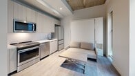 Located at 5522 Baum Blvd in Pittsburgh's Shadyside neighborhood, this efficiently designed studio maximizes every inch of its compact footprint across a single floor. The open-plan kitchen and living area serves as the heart of the space, showcasing exposed concrete ceiling beams that lend an industrial character alongside clean white walls and light wood-tone flooring. The kitchen is fully equipped with stainless steel appliances — including a French-door refrigerator, dishwasher, range, and built-in microwave — complemented by light wood cabinetry extending to the ceiling for ample storage and a sleek white countertop. The living area is thoughtfully furnished with a wall-mounted Murphy bed system integrated with a sofa and storage shelving, allowing the room to function as both a lounge and sleeping space. A separate hallway leads to a dedicated utility room housing a stacked washer and dryer. The hallway also connects to a workspace area with a desk, wall-mounted TV console, and additional storage cabinetry. The bathroom features clean-lined tile flooring, a glass-enclosed walk-in shower, and modern fixtures. Natural light filters in through windows, brightening the neutral palette throughout.