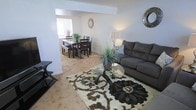 This two-floor residence offers a welcoming and practical layout with a seamlessly connected living and dining area as a central feature. The living space is anchored by plush wall-to-wall carpeting in a warm neutral tone, complemented by a decorative patterned area rug in earthy browns and creams. A comfortable sectional-style sofa in charcoal gray provides ample seating, while a round glass-top coffee table adds a contemporary touch. Decorative greenery and wall accents contribute to the room's inviting ambiance.

An open passage connects the living area to the dining space, which features a dark-stained dining table with matching chairs and a bench, set atop a coordinating area rug. Natural light filters through a window at the far end of the dining room, brightening the space and enhancing its open feel. The consistent neutral wall tones and light carpeting throughout create a cohesive, airy atmosphere. The two-floor configuration suggests additional living or sleeping quarters above, offering a functional separation between communal and private areas of the home.