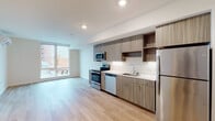 This thoughtfully designed single-floor unit offers a clean, contemporary aesthetic throughout, characterized by light wood-tone flooring, crisp white walls, and recessed lighting that enhance the bright, airy atmosphere.

The open-concept kitchen and living area forms the heart of the unit. The kitchen is outfitted with wood-grain cabinetry featuring a two-tone arrangement of upper and lower cabinets, complemented by open shelving for added display and storage. Gleaming white countertops provide ample workspace, while a full suite of stainless steel appliances — including a range, microwave, dishwasher, and refrigerator — underscores the modern finish. Under-cabinet lighting adds a polished touch. The kitchen flows seamlessly into an open living area, which benefits from a large window that draws in natural light and frames views of the surrounding urban streetscape.

The bedroom is a standout space, featuring expansive floor-to-ceiling windows that flood the room with daylight and offer open city views. Two sliding-door closets provide generous built-in storage, keeping the room clean and functional.

The bathroom is finished with the same warm wood-tone vanity cabinetry seen throughout the unit, paired with a white countertop and a full tub-shower combination. Light wood flooring extends into this space, maintaining a cohesive design language across the entire unit.