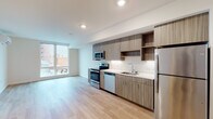 This thoughtfully designed single-floor unit offers a clean, contemporary aesthetic throughout, characterized by light wood-tone flooring, crisp white walls, and recessed lighting that enhance the bright, airy atmosphere.

The open-concept kitchen and living area forms the heart of the unit. The kitchen is outfitted with wood-grain cabinetry featuring a two-tone arrangement of upper and lower cabinets, complemented by open shelving for added display and storage. Gleaming white countertops provide ample workspace, while a full suite of stainless steel appliances — including a range, microwave, dishwasher, and refrigerator — underscores the modern finish. Under-cabinet lighting adds a polished touch. The kitchen flows seamlessly into an open living area, which benefits from a large window that draws in natural light and frames views of the surrounding urban streetscape.

The bedroom is a standout space, featuring expansive floor-to-ceiling windows that flood the room with daylight and offer open city views. Two sliding-door closets provide generous built-in storage, keeping the room clean and functional.

The bathroom is finished with the same warm wood-tone vanity cabinetry seen throughout the unit, paired with a white countertop and a full tub-shower combination. Light wood flooring extends into this space, maintaining a cohesive design language across the entire unit.