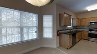 This two-story space presents a clean, functional layout with warm neutral tones and practical design details. The main level showcases an open area where the kitchen flows seamlessly into the living space. The kitchen is equipped with light wood cabinetry, dark granite-style countertops, and stainless steel appliances, offering a balanced contrast between warm and modern finishes. A single-basin sink sits beneath a window, allowing natural light to brighten the workspace.

Two generously sized windows fitted with white horizontal blinds line the adjacent wall, drawing in ample natural light and providing views of surrounding greenery. The walls are finished in a warm taupe tone, complementing the light carpeted flooring that extends through the living area. White baseboards add a crisp, finished look throughout.

The vaulted or angled ceiling detail adds a sense of height and openness to the space. A fire extinguisher is mounted near the kitchen, noting attention to safety. The overall ambiance is understated and functional, with a neutral palette that offers flexibility for personalization across both floors.