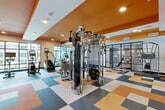 Located at 2801 Windy Ridge Parkway SE in Atlanta, Georgia, this well-appointed fitness center offers a generously sized, single-floor facility designed to support a comprehensive range of workout routines. The space is immediately distinguished by its bold interior design, featuring striking orange accent ceilings and walls that contrast with clean white surfaces, creating an energetic and motivating atmosphere throughout.

The flooring transitions across two distinct zones: a vibrant multi-colored checkerboard tile area that anchors the cardio and cable machine sections, and a warm wood-look hardwood floor that defines the free weights and functional training area. Large floor-to-ceiling windows line the perimeter, flooding the space with natural light and providing open views of the surrounding Windy Ridge area.

The cardio zone is equipped with multiple treadmills, ellipticals, stair climbers, and stationary bikes, many featuring integrated digital displays. A centrally positioned cable crossover machine serves as a focal point of the strength training area, complemented by a full rack of selectorized weight machines. The free weights section offers barbells, a squat rack, weight benches, stability balls, and plyo boxes for functional fitness. A ceiling-mounted fan, recessed lighting, and wall-mounted television screens enhance comfort and usability. Glass-paneled interior partitions add a modern architectural touch, while clearly marked entry and exit doors ensure easy access throughout.