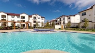 This expansive outdoor pool area is designed to deliver a resort-style experience. At its center is a generously sized swimming pool with crystal-clear blue water, complemented by a raised spa and a shallow sun shelf — ideal for relaxing at the water's edge. The pool deck is finished in textured concrete and surrounded by a row of contemporary chaise lounges, providing ample space for sunbathing. Tall palm trees are strategically placed around the deck, reinforcing the tropical ambiance against an open sky.

Adjacent to the pool is a covered pavilion that serves as a shaded retreat. The structure is supported by substantial natural stone columns and features a warm wood-paneled ceiling, ceiling fans, and large tile flooring in a neutral stone finish. Glass railings along the pavilion edge maintain unobstructed sightlines to the pool. Modern woven seating is positioned throughout the covered area, offering a comfortable lounge space out of direct sun.

The entire pool area is enclosed by black metal fencing, providing security while preserving the open, airy feel of the space. The surrounding residential buildings, finished in white stucco with red tile rooflines, frame the environment and contribute to a cohesive Mediterranean-inspired aesthetic throughout the community.