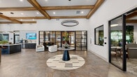 Located at 5000 Collina Ter in Clermont, Florida, this beautifully appointed single-floor community clubhouse offers a generously sized gathering space designed with upscale finishes and a warm, modern aesthetic. The interior is anchored by striking exposed wooden beam ceilings that soar overhead, complemented by crisp white walls and durable stone-tile flooring throughout.

The welcoming entrance foyer features a dramatic circular pendant light and a central marble-topped accent table, setting an elegant tone upon arrival. Black-framed glass partition walls divide the space with visual openness, allowing natural light to flow freely between zones.

A focal point of the lounge area is a floor-to-ceiling stacked-stone fireplace, flanked by built-in seating and topped with a large flat-screen display — creating a cozy yet refined gathering spot. Adjacent seating areas are furnished with contemporary sofas and accent chairs arranged for both conversation and relaxation.

The entertainment zone includes a shuffleboard table, additional lounge seating, and a stylish wet bar with counter seating and a decorative feature wall. Large windows throughout offer views of the surrounding outdoor areas, filling the space with abundant natural light. The cohesive design blends rustic and contemporary elements, resulting in a sophisticated and inviting atmosphere ideal for community gatherings.