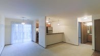 This single-floor space is designed with an open-concept layout that connects the living area and kitchen, creating a cohesive and functional floor plan. The living area is finished with neutral carpet and light-toned walls, providing a clean and versatile backdrop. A half-wall peninsula with a dark countertop separates the kitchen from the living space, offering both a visual divider and a practical surface for dining or food preparation. The kitchen is equipped with stainless steel appliances, including a refrigerator and a range, and features warm wood-toned cabinetry that adds a touch of contrast to the otherwise neutral palette. Three pendant lights hang above the peninsula, providing focused task lighting while enhancing the overall ambiance. A sliding glass door at the far end of the space allows natural light to flow in and provides direct access to an outdoor patio area, further brightening the interior. The layout is efficient and well-organized, with clear sightlines throughout, making the space feel open and airy despite its compact footprint.