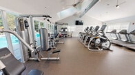 This fitness center offers a generously sized, single-level workout space designed to accommodate a comprehensive range of exercise needs. The room is defined by its striking vaulted ceiling, which rises dramatically overhead and is fitted with skylights that flood the space with natural light. An exposed metal ductwork system runs along the ceiling, adding an industrial accent to the otherwise clean, neutral interior palette of white and soft gray walls.

The durable, speckled rubber flooring spans the entire area, providing a safe and resilient surface suitable for all workout activities. Ceiling fans are strategically positioned throughout to ensure consistent air circulation. Multiple wall-mounted flat-screen televisions are placed at convenient viewing angles across the space.

The cardio section is well-appointed with a row of elliptical trainers and several Matrix treadmills, while the strength training area features a multi-station cable machine, a dumbbell rack stocked with a full range of weights, and various benches and free weight stations. Large windows fitted with white blinds line the walls, allowing natural light to enter while offering privacy. A glazed door and windows on one side provide views of an outdoor pool area, connecting the interior to the surrounding outdoor amenity space.