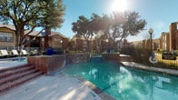 This single-story property offers a well-appointed communal outdoor area centered around a generously sized swimming pool with clearly marked depth zones. The pool area is framed by classic brick coping and raised planters accented with bold blue ceramic pots, adding a touch of color to the sunlit courtyard. Mature oak trees provide natural shade throughout the space, while a wrought-iron perimeter fence encloses the area for privacy and security. Lounge seating and a covered pavilion are visible nearby, offering additional outdoor gathering options beneath the open Texas sky.

The interior space features a vaulted ceiling with dual ceiling fans, creating an airy, open atmosphere in the main living area. Recessed lighting complements the natural light that filters through multiple windows. The kitchen is finished with granite countertops in warm golden tones, dark espresso cabinetry, and a decorative mosaic tile backsplash in muted blues and browns. Black appliances, including a refrigerator, range, built-in microwave, and dishwasher, complete the kitchen. A double stainless steel sink and a pass-through counter connect the kitchen to the open living and dining area, making the overall layout feel cohesive and functional.