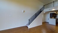 This space showcases a thoughtfully designed interior highlighted by a striking staircase with ornate wrought iron balusters featuring decorative scroll detailing and a clean white stringer. The railing's dark finish provides a sophisticated contrast against the neutral, light-toned walls, creating a visually appealing focal point upon entry. The flooring throughout is warm hardwood in a natural tone, lending the space a classic and inviting feel. The walls are finished in a soft, neutral palette that maximizes the sense of brightness and openness. A subtle wall sconce adds a refined lighting accent, contributing to the overall polished atmosphere. The space beneath and around the staircase is left open, enhancing the sense of spaciousness. An arched doorway is visible in the background, hinting at architectural detailing that adds character to the layout. The clean lines, neutral tones, and decorative ironwork combine to create an environment that feels both elegant and welcoming, suitable for a variety of interior styles and furnishing choices.